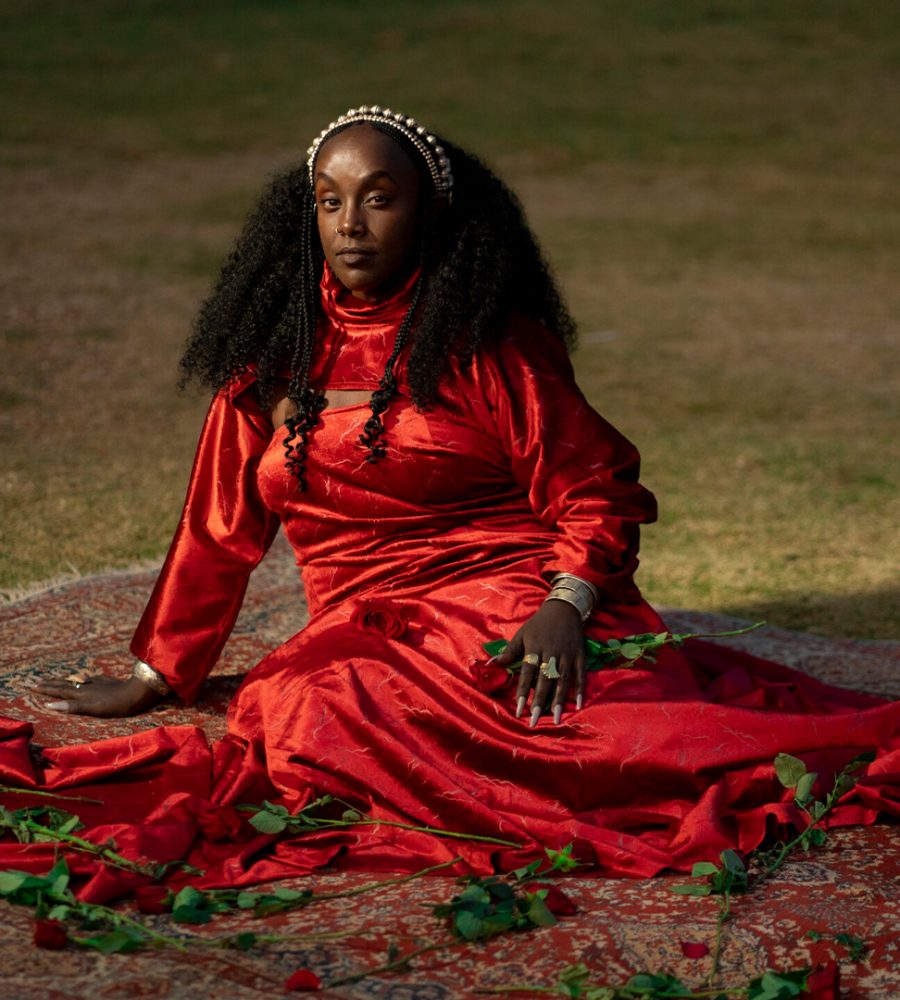 En kvinna i röd klänning sittandes på en röd matta i gräset omgiven av röda rosor